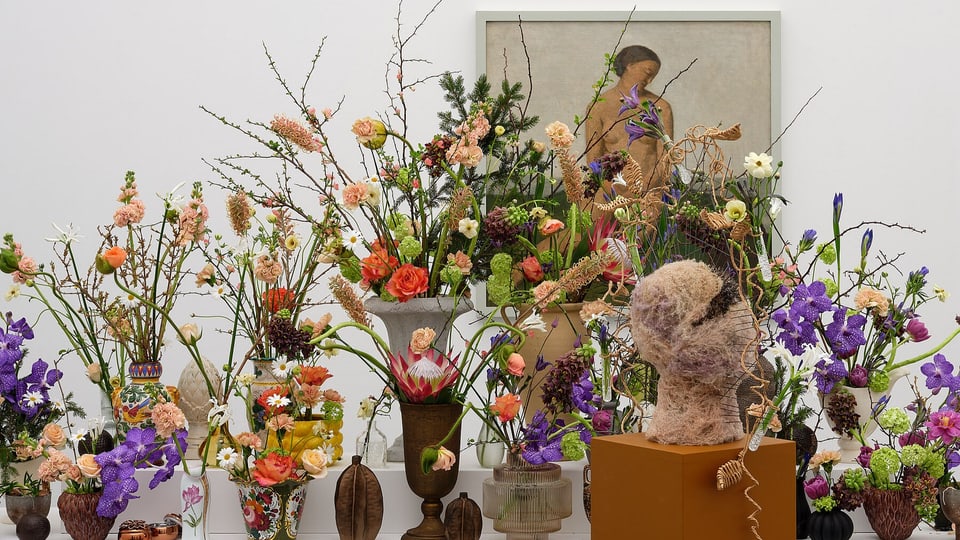 Das Gemälde einer nackten Frau, davor bunte Blumen in verschiedenen Vasen. 