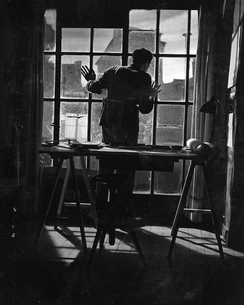Ein Mann steht mit beiden Händen gegen eine Fensterfront gelehnt und blickt hinaus.