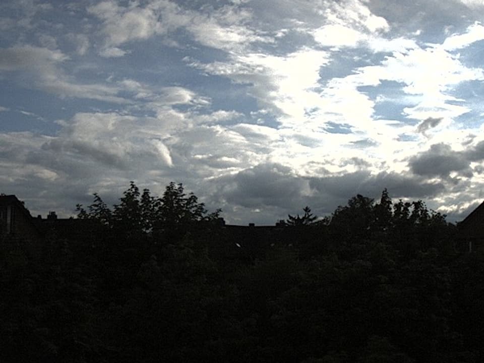 Grösstenteils ist ein wechselnd bewölkter Himmel mit blauen Flecken und weissen und grauen Wolken zu sehen. Am unteren Rand des Bildes sind die dunklen Konturen von Büschen und Dächern zu sehen.