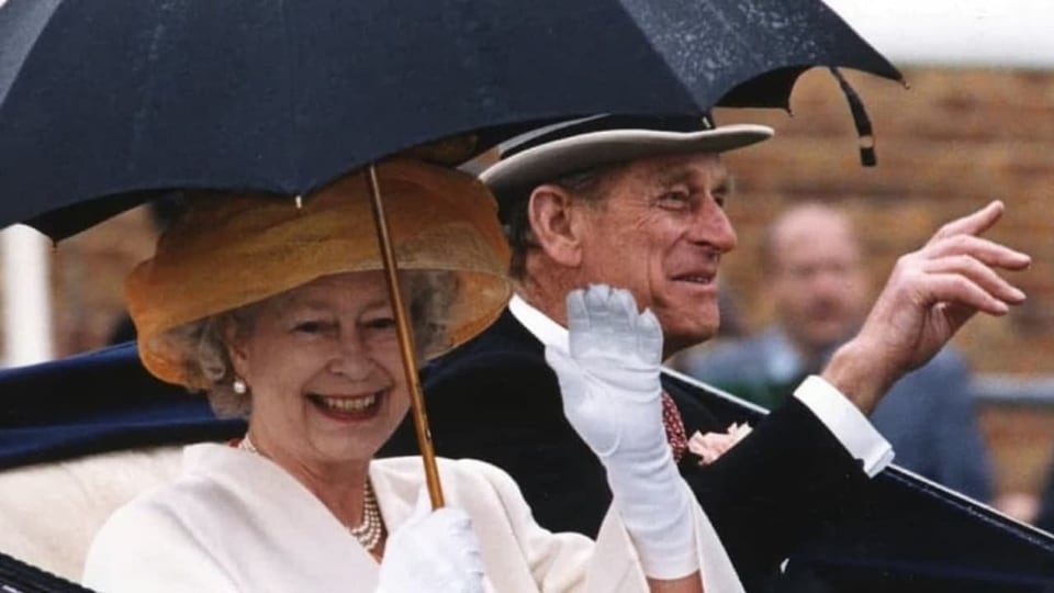 Die Queen und ihr Mann Philip fahren in einer Kutsche durch den Regen, geschützt von einem schwarzen Schirm. 