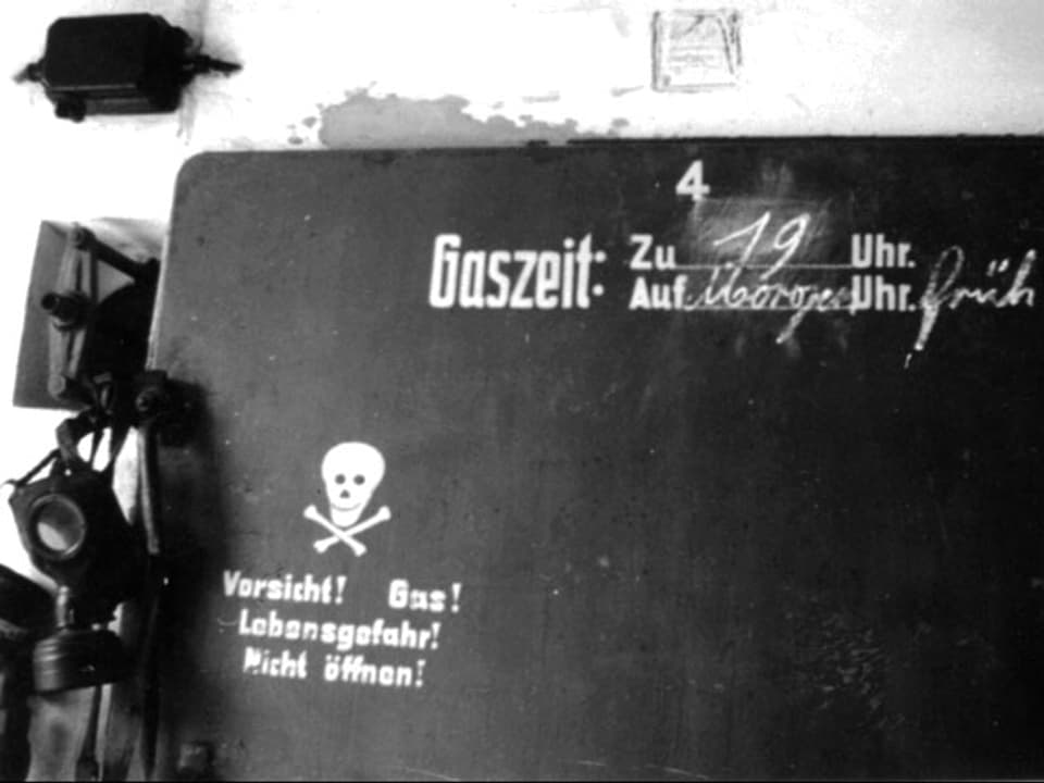 Eine Gasmaske hängt auf einer Schiefertafel. Auf der Tafel ist ein lächelnder Totenkopf abgebildet und die «Gaszeit» notiert.