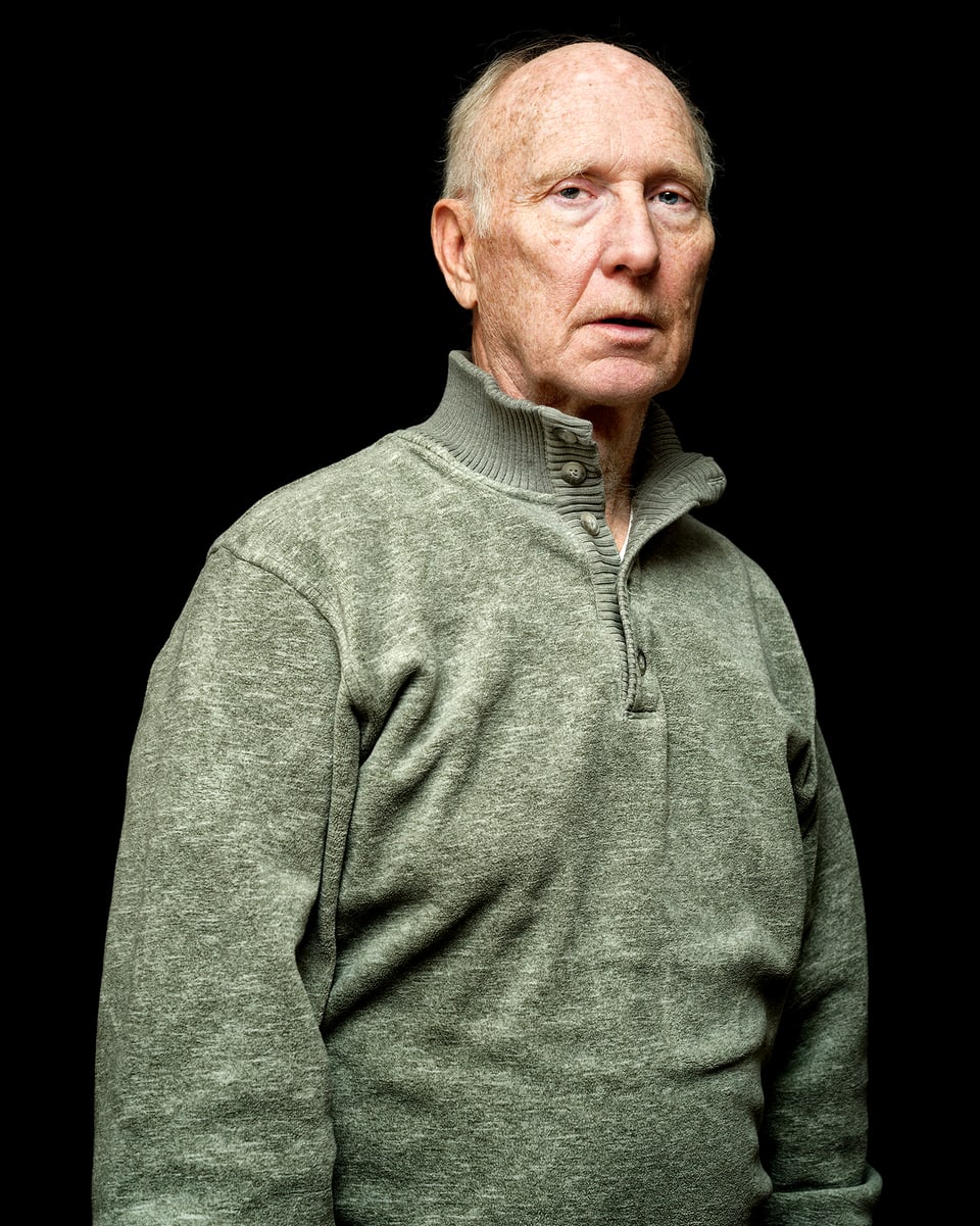Ein älterer Mann in grau-grünem Pullover vor schwarzem Hintergrund.