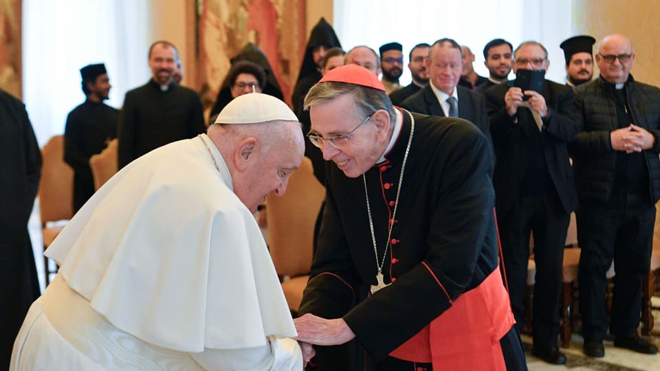 Mann in weiss und weiterer Mann in Kardinalskutte schütteln Hände.