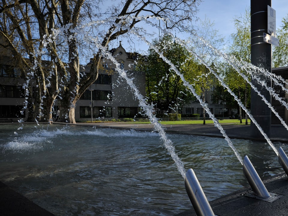 Im Vordergrund ist ein Springbrunnen in einem Park zu sehen, im Hintergrund das Dreirosen-Schulhaus.
