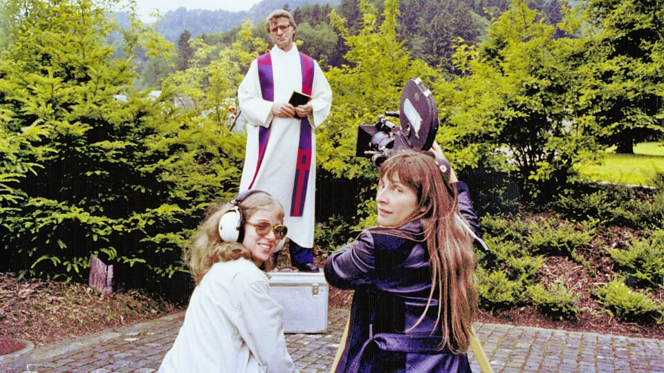 Drei Personen schauen in die Kamera. Zwei Frauen, eine hantiert an der Kamera die andere hat Kopfhörer auf. Sie sind auf einem Filmset, im hintergrund ist ein Pfarrer zu sehen.