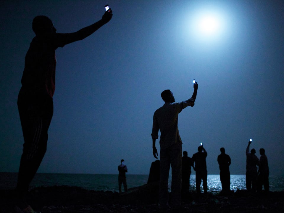 Menschen halten in der Nacht und am Meeresrand ihre Smartphones in die Höhe, die Displays leuchten hell.