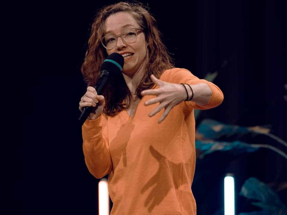Martina Hügi gestikuliert auf der Bühne mit orangem Pullover.