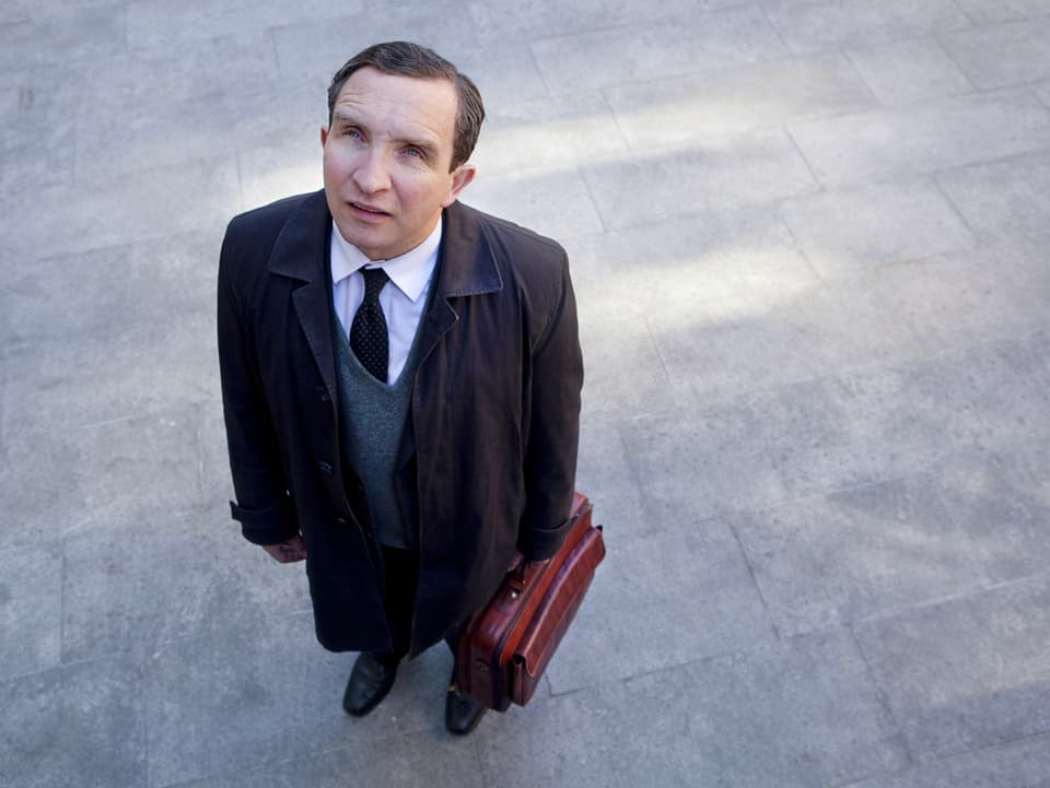 Ein Mann mittleren Alters mit Krawatte und Aktentasche in der Hand steht auf einem Platz und guckt nachoben.