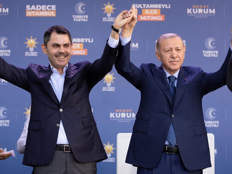Erdogan und Kurum stehen gemeinsam auf einer Bühne und halten zusammen die Hände in die Luft.