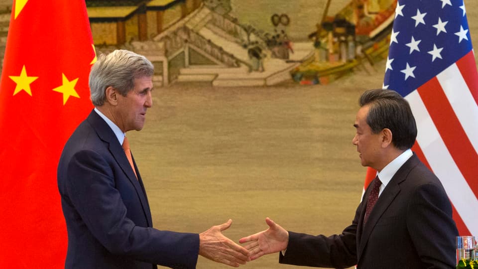 John Kerry und Wang Yi strecken sich die Hände entgegen, im Hintergrund eine chiesische und eine amerikanische Fahne
