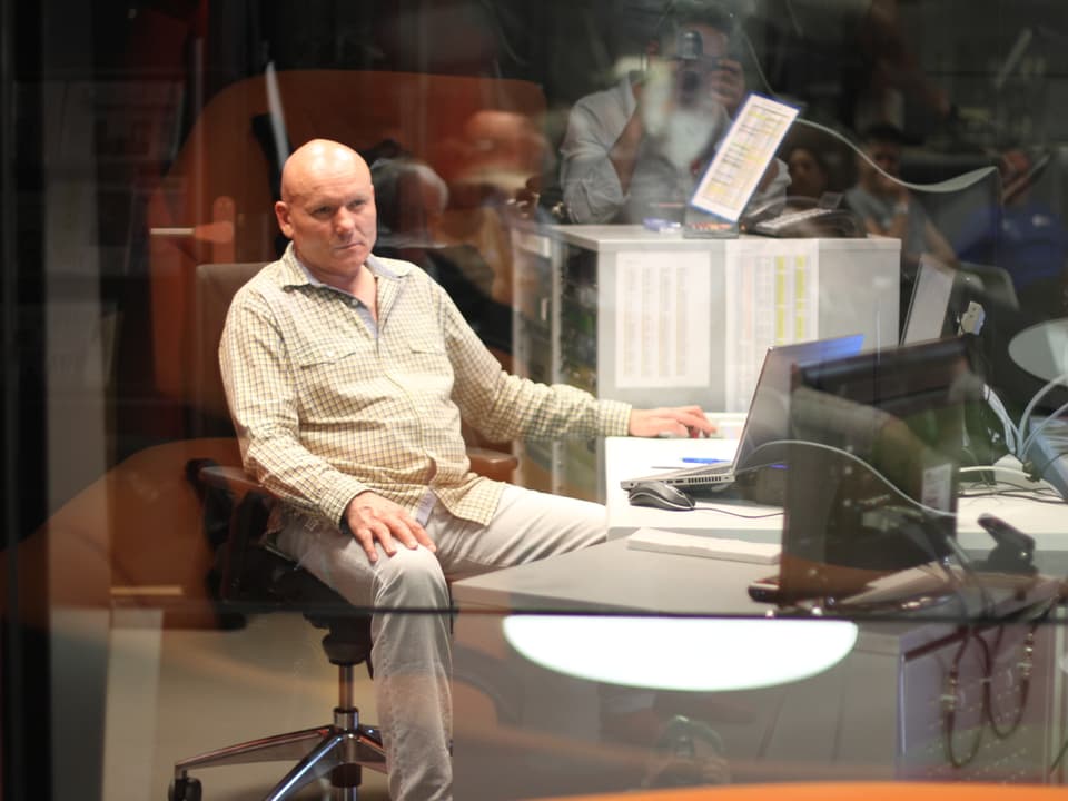 Mann sitzt hinter Glas in einem Technikraum.