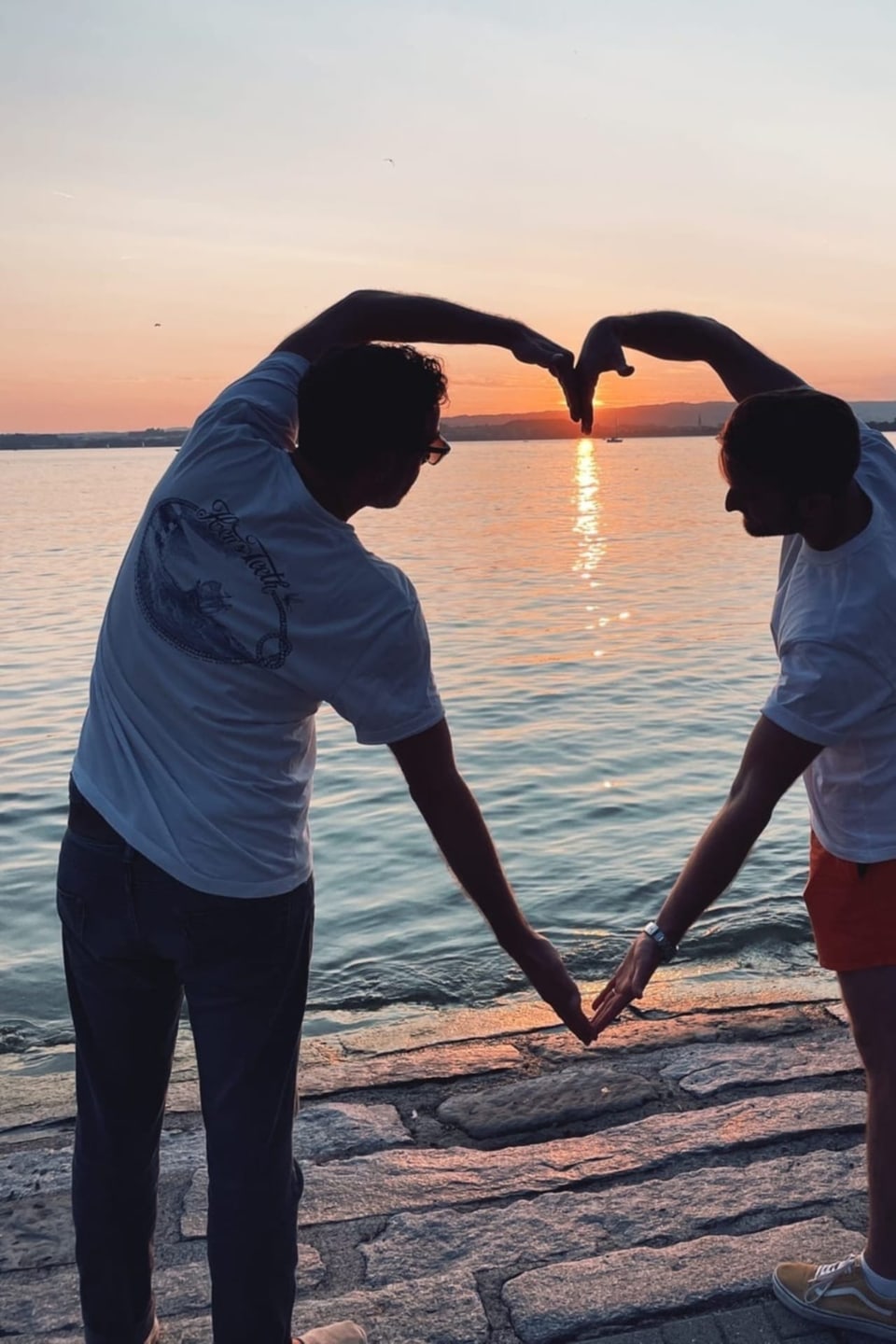 Zwei Männer am Ufer eines Sees bei Sonnenuntergang formen ein Herz mit ihren Armen.