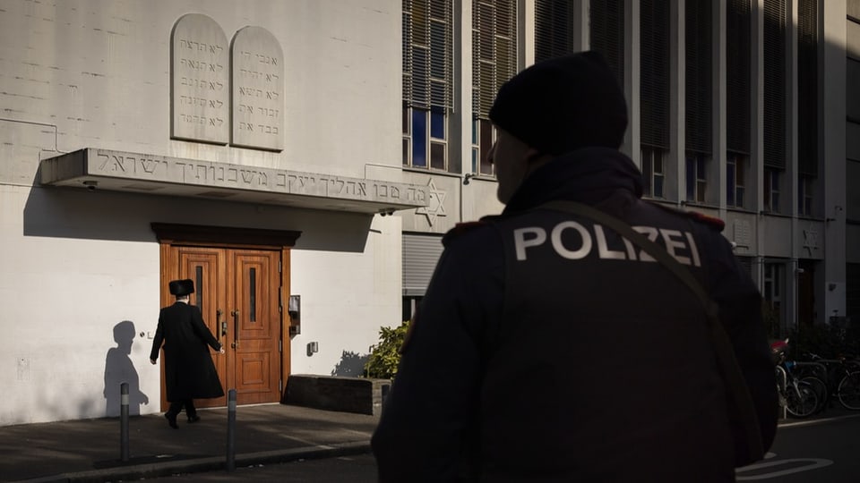 Ein orthodoxer Jude mit schwarzer Bekleidung vor dem Eingang zur Synagoge. Ein Polizist steht in der Nähe.