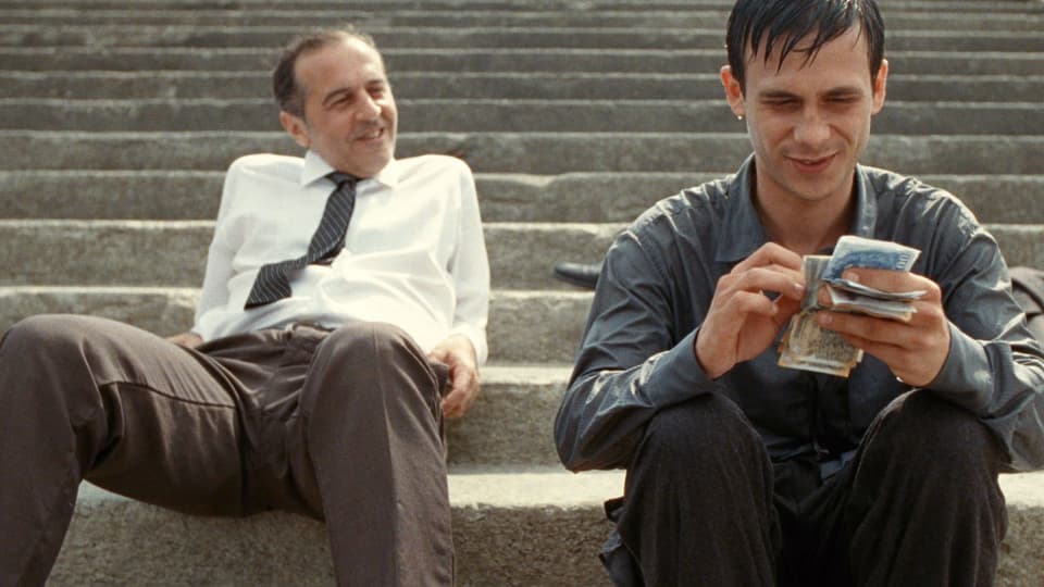 Zwei Männer sitzen auf einer Treppe. Ein älterer mit Hemd und Krawatte lehnt sich lässig nach hinten. Ein jüngerer zählt lächelnd Geldscheine.