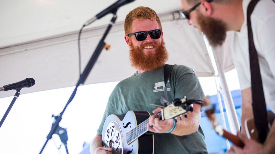 Mann mit Gitarre, Sonnenbrille und T-Shirt musiziert lächelnd auf einer Bühne neben einem Kollegen