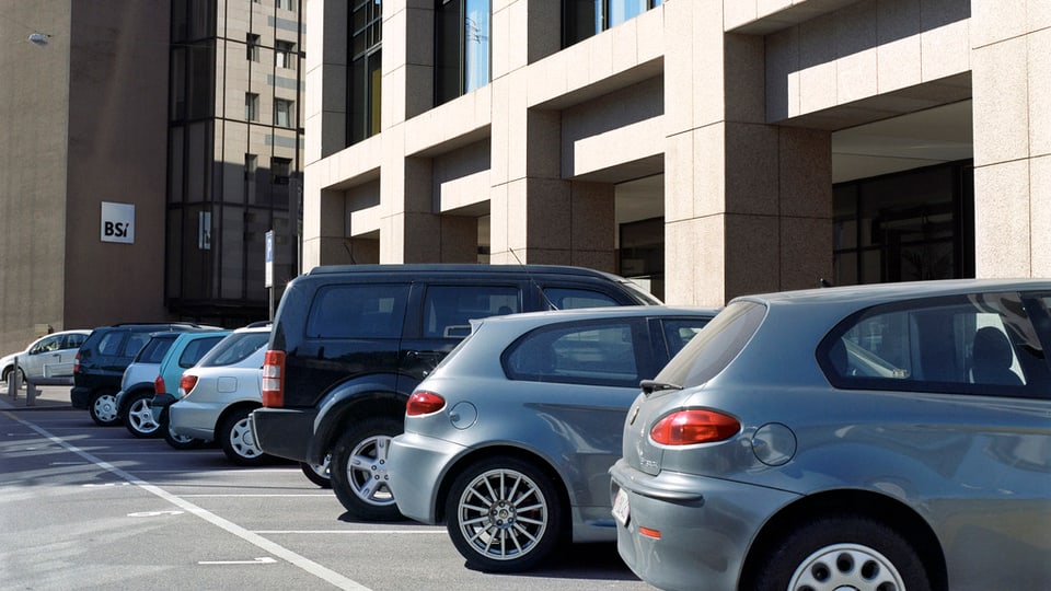 Einige schräg einparkierte Autos vor einem modernen Gebäude, im Hintergrund ein zweiter Neubau.