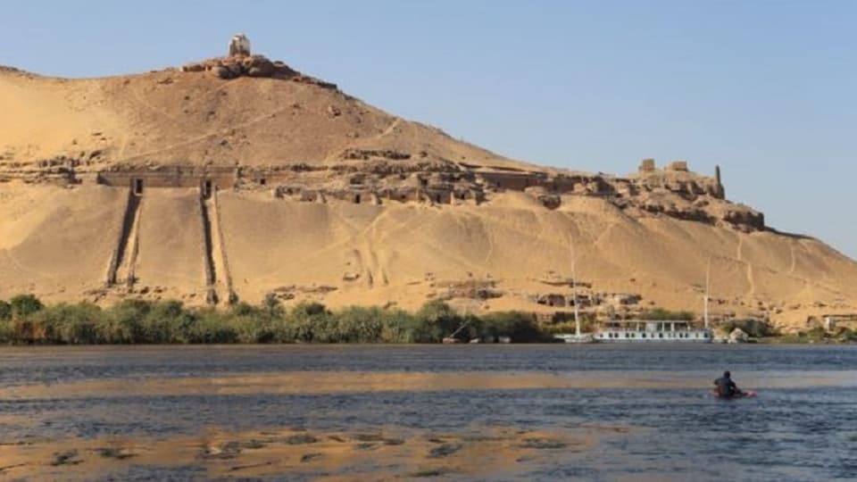 Auf dem Bild sieht man der Nil, im Hintergrund ein brauner Hügel.