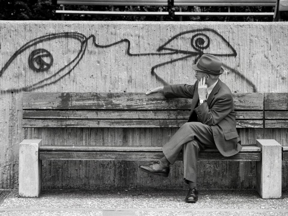 Ein Mann sitzt auf einer Bank und dreht sich um zu einer Zeichnung von Harald Naegeli an der Wand hinter ihm.
