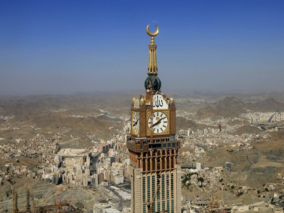 Ein riesiger Turm mit Turmuhr und goldener Mondsichel steht unter blauem Himmel inmiten einer Stadt, die in der Wüste steht.