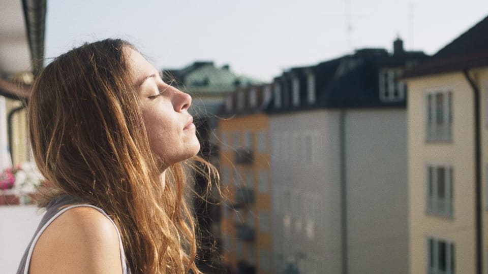 Eine Frau steht auf dem Balkon und richtet ihr Gesicht mit geschlossenen Augen zur Sonne.