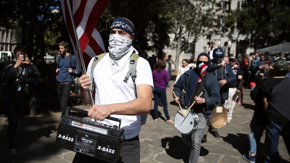 Ein maskierter Mann trägt Amerikaflagge auf einer Demonstration.