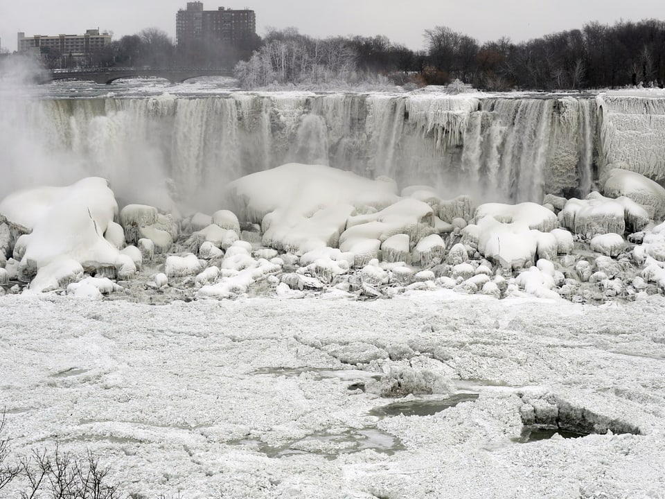 Die Niagarafälle sind mit Eisbildungen dekoriert, im Hintergrund sind Häuser zu sehen.