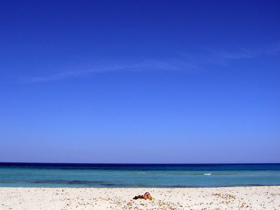 Zu sehen ist im unteren Drittel der Fotografie ein Stück Sandstrand mit einem Sonnenbadenden, der die Füsse zum Fotografen hin hält, und ein Stück Meer. Die oberen zwei Drittel der Fotografie nimmt ein blauer Himmel ein.