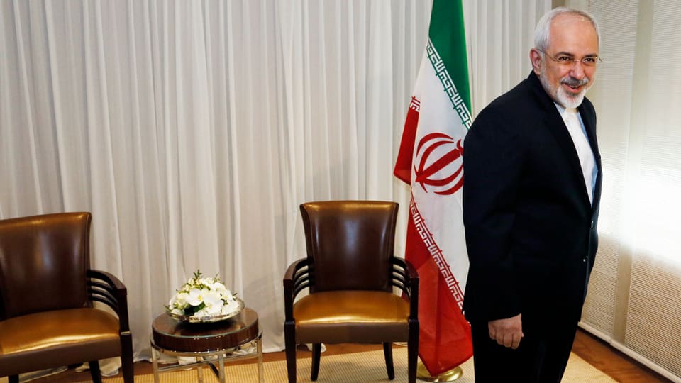 Irans Aussenminister Sarif stehend vor zwei Sesseln und einer iranischen Fahne.