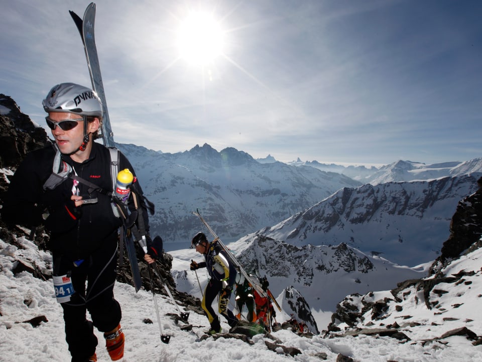 Alpenpanorama bei Sonneschein. Verschwitzte Tourenläufer kämpfen sich den Berg hoch, die Skier aber auf dem Rücken. 