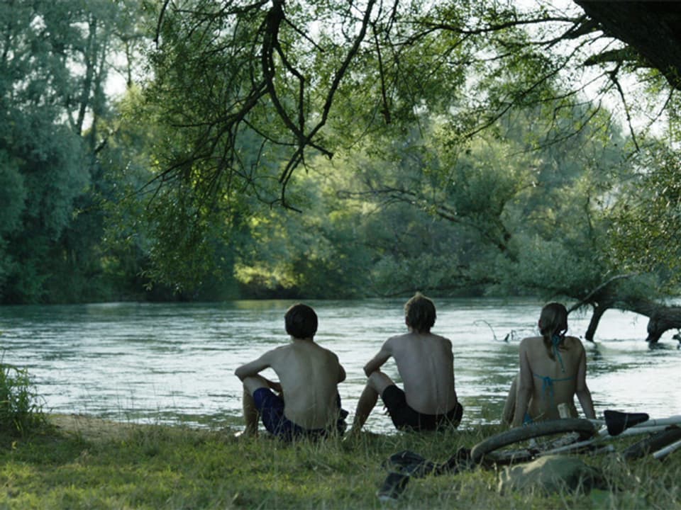Drei Jugendliche sitzen am Flussufer und schauen zum Wasser. 