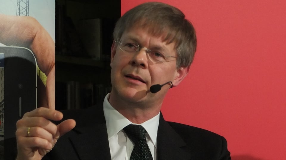 Gerhard Schafroth argumentiert auf einem Wahlpodium und deutet mit Daumen und Zeigfinder eine Distanz an.