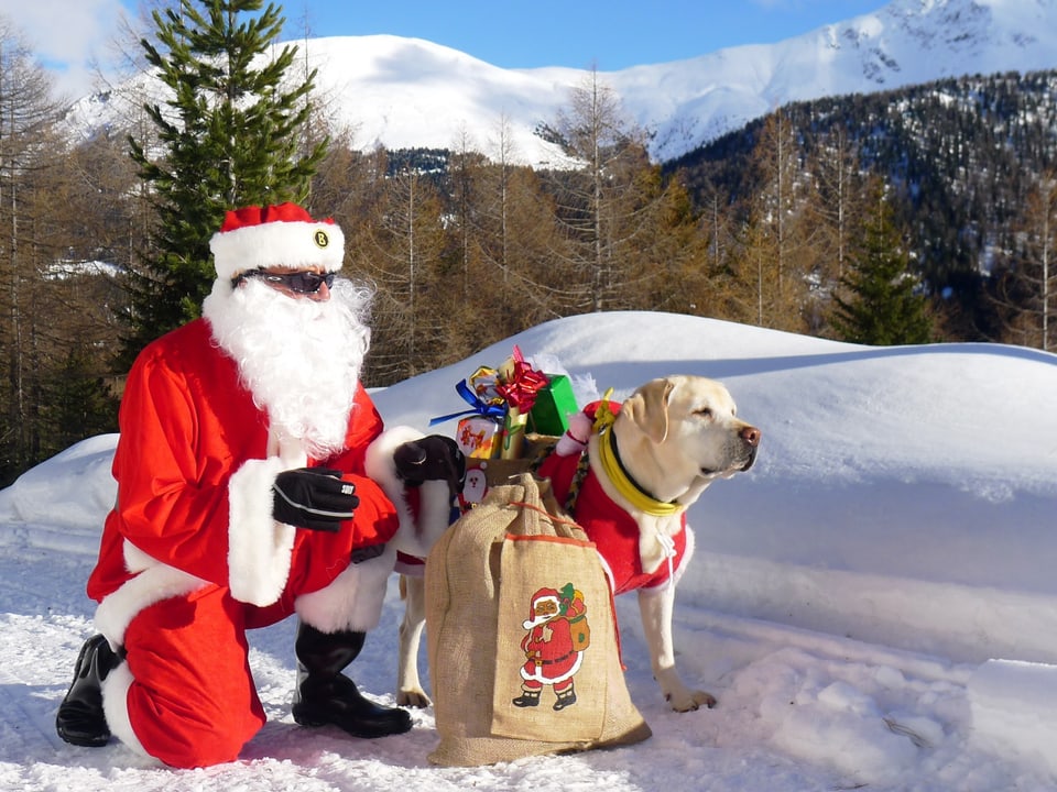 Ein Nikolaus im roten Dress kniet im Schnee vor einem Sennhund der Geschenke trägt, dahinter verschneite Graubündner Berge.