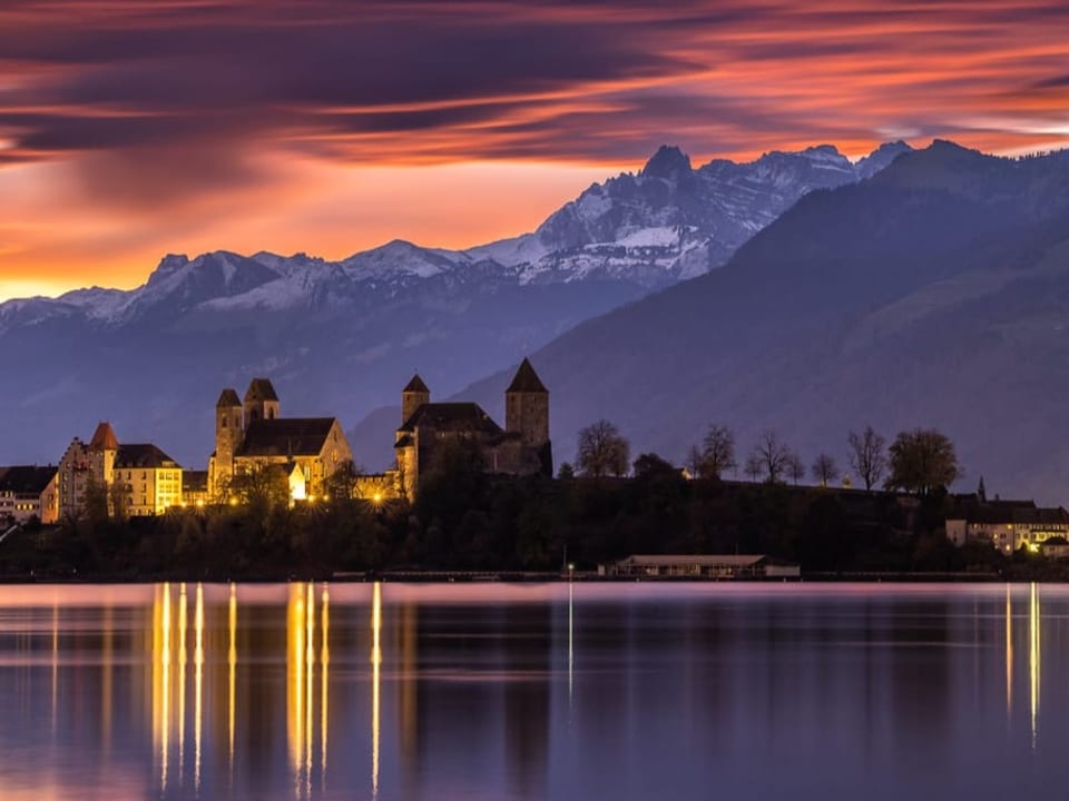 Blick über den See auf Altstadt, im Hintergrund Alpen mit rot leuchtenden Wolken.