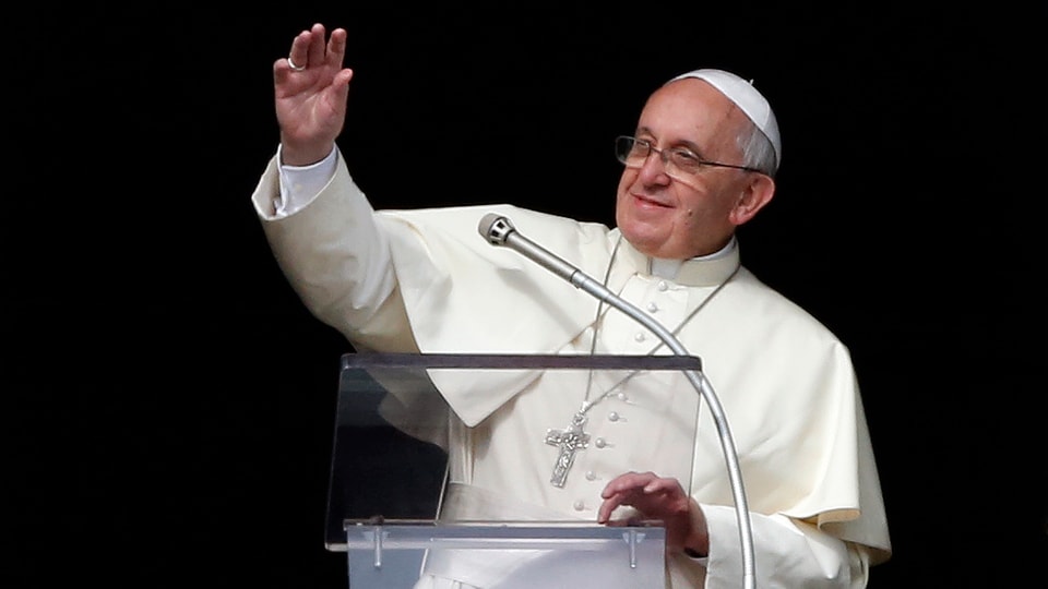 Papst Franziskus winkt vor schwarzem Hintergrund der Menge zu.