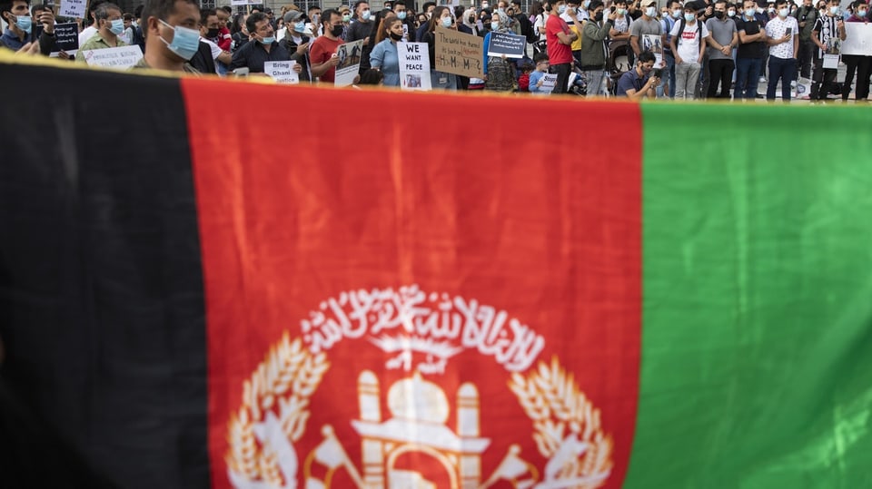 Hochgehaltene Flagge Afghanistans; im Hintergrund sind demonstrierende Personen zu sehen