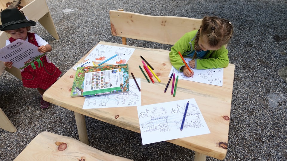 Ein Kind sitzt an einem kleinen Holztisch und malt mit einem Farbstift Tiere aus. 