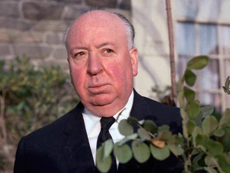 Alfred Hitchcock blickt hinter einem Strauch hervor.