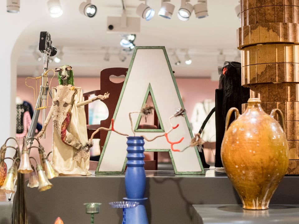 Ein Leuchtbuchstaben A neben einer Vase, Lampe und Skulpturen.