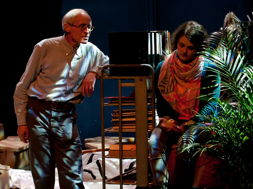 Martin Vosseler steht auf der Theaterbühne. Aninna Poliva sitzt auf einem Stuhl, inmitten von Bühnenrequisiten.