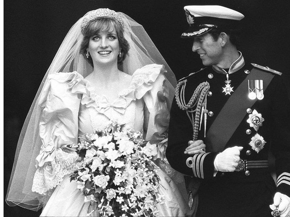 Lady Di lächelt am Arm ihres Gatten im Hochzeitskleid in die Kamera.