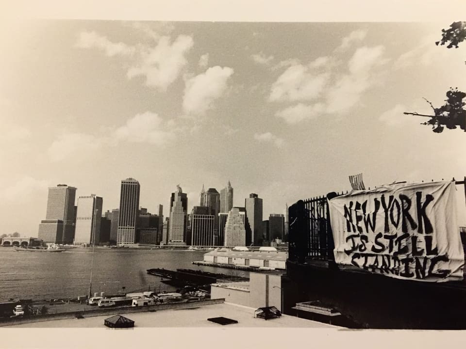 Auf dem Bild ist ein Transparent zu sehen, auf dem steht: New York is still standing. Im Hintergrund die Skyline.