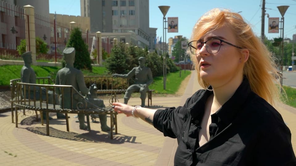 Frau in schwarzer Bluse zeigt auf Statue aus Bronze hinter sich