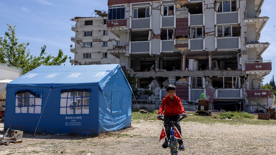 Ein Junge fährt auf einem Velo, neben ihm ist ein provisorisches Zelt und dahinter ein zerstörtes Wohnhaus zu sehen