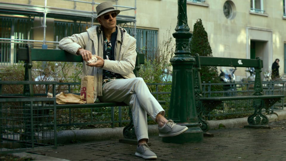 Michael Fassbender sits on a park bench as a well-masked killer wearing a hat and sunglasses.