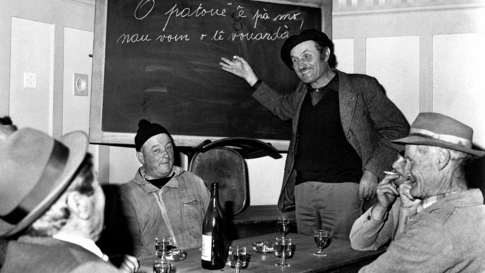 Schwarzweissfoto: Männer vor Weingläsern an einem Tisch, dahinter eine Wandtafel mit handschriftlichem Text