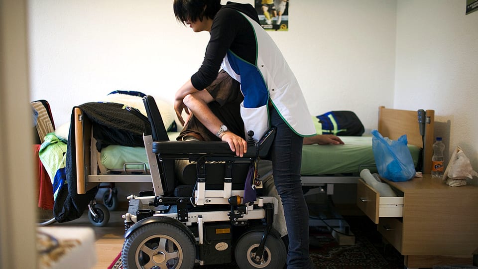 Eine Pflegerin hilft einem Behinderten in seinen Rollstuhl.