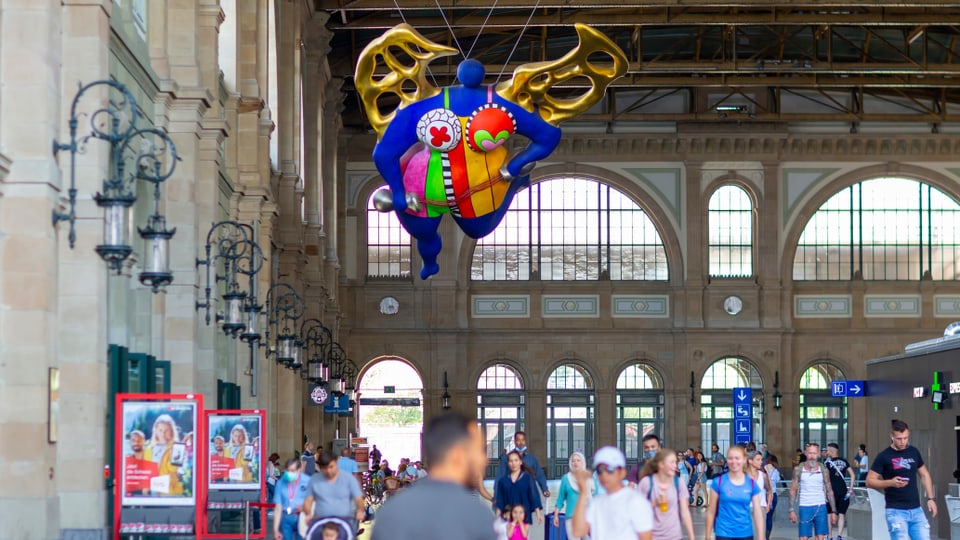 Ein bunter Engel in einer vollen Bahnhofshalle.
