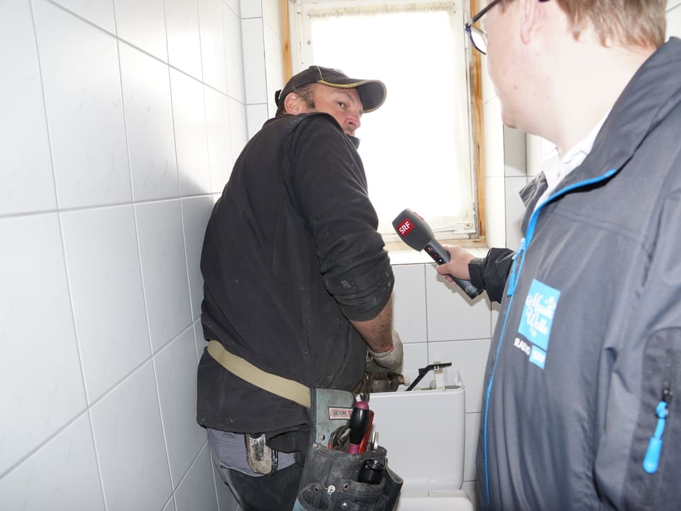 Der Reporter befragt einen Handwerker, die auf einer Toilette mit dem Spülkasten beschäftigt ist.
