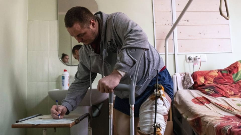 Ein Verwundeter unterschreibt einen Zettel neben dem Spitalbett