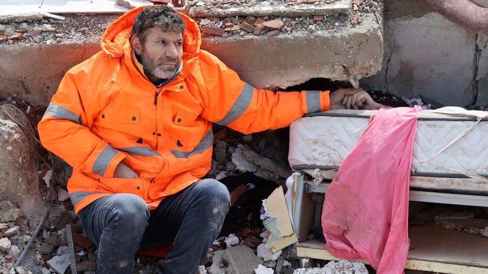 Ein Mann in einer orangen Jacke hält eine Hand, die aus den Trümmern ragt